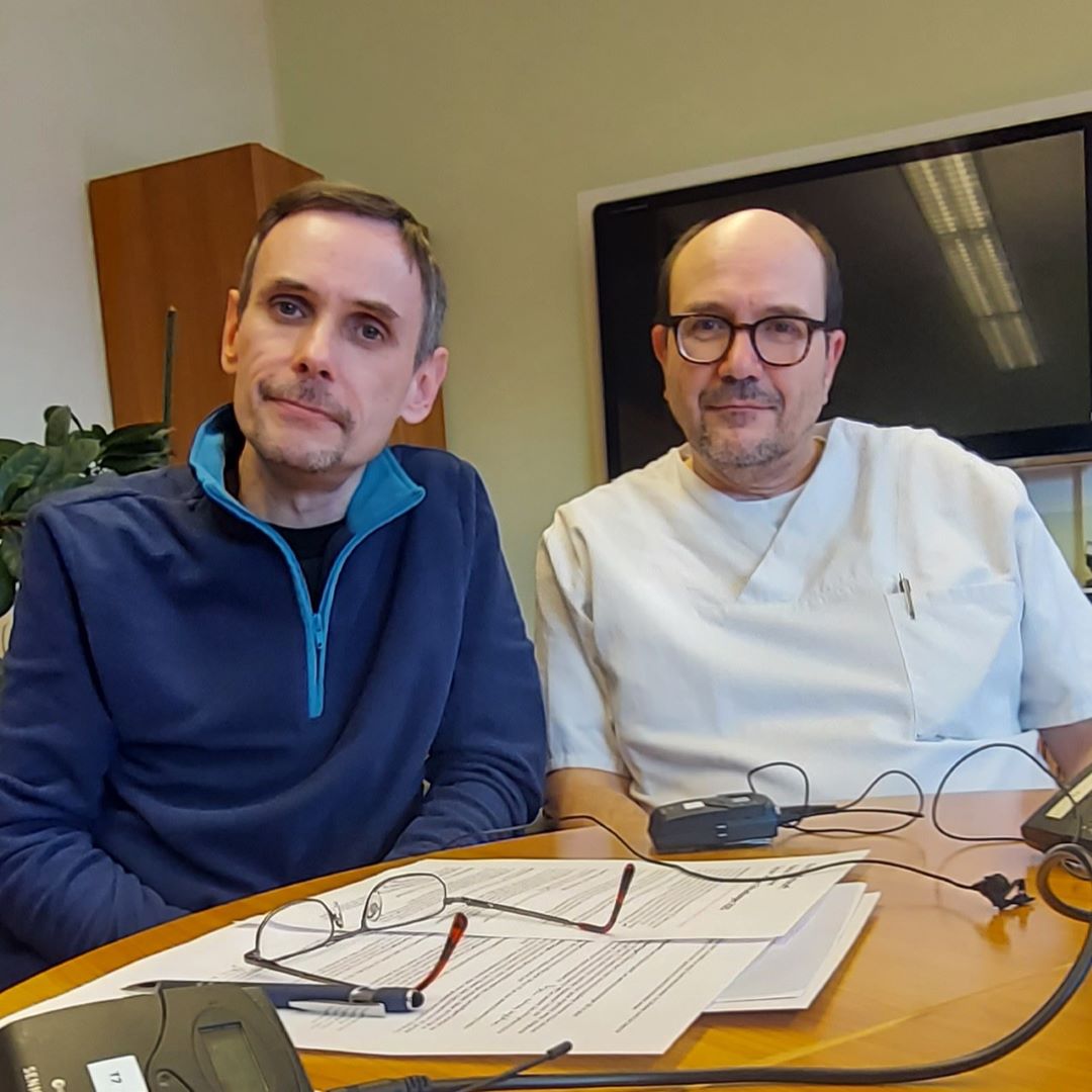 Michael Brumme und Dr. med. Albert Grüger sitzen sich an einem Tisch und sprechen in einem Interview über Cluster-Kopfschmerzen. Auf dem Tisch liegen Dokumente, Stifte und Mikrofone.